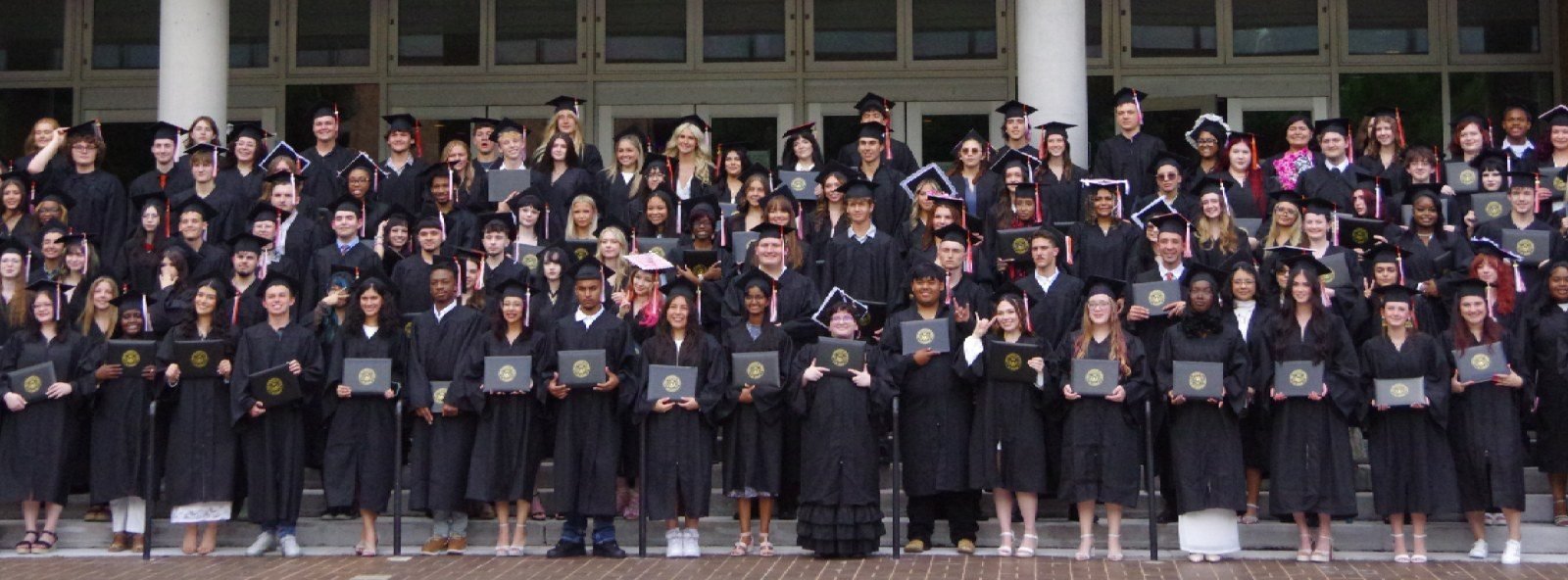 UT High School graduates in black caps and gowns holding their diplomas on the steps of a campus building